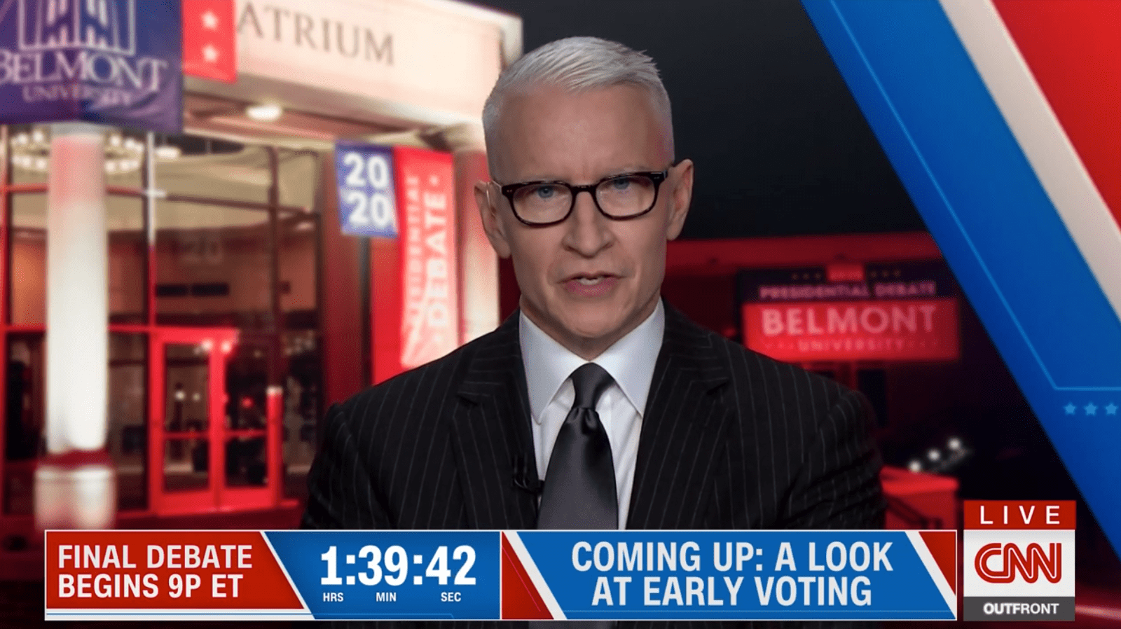 A man, Anderson Cooper, reports for CNN in front of Belmont's Maddox Grand Atrium, illuminated with red and blue lights and decorated with Belmont and Debate posters.