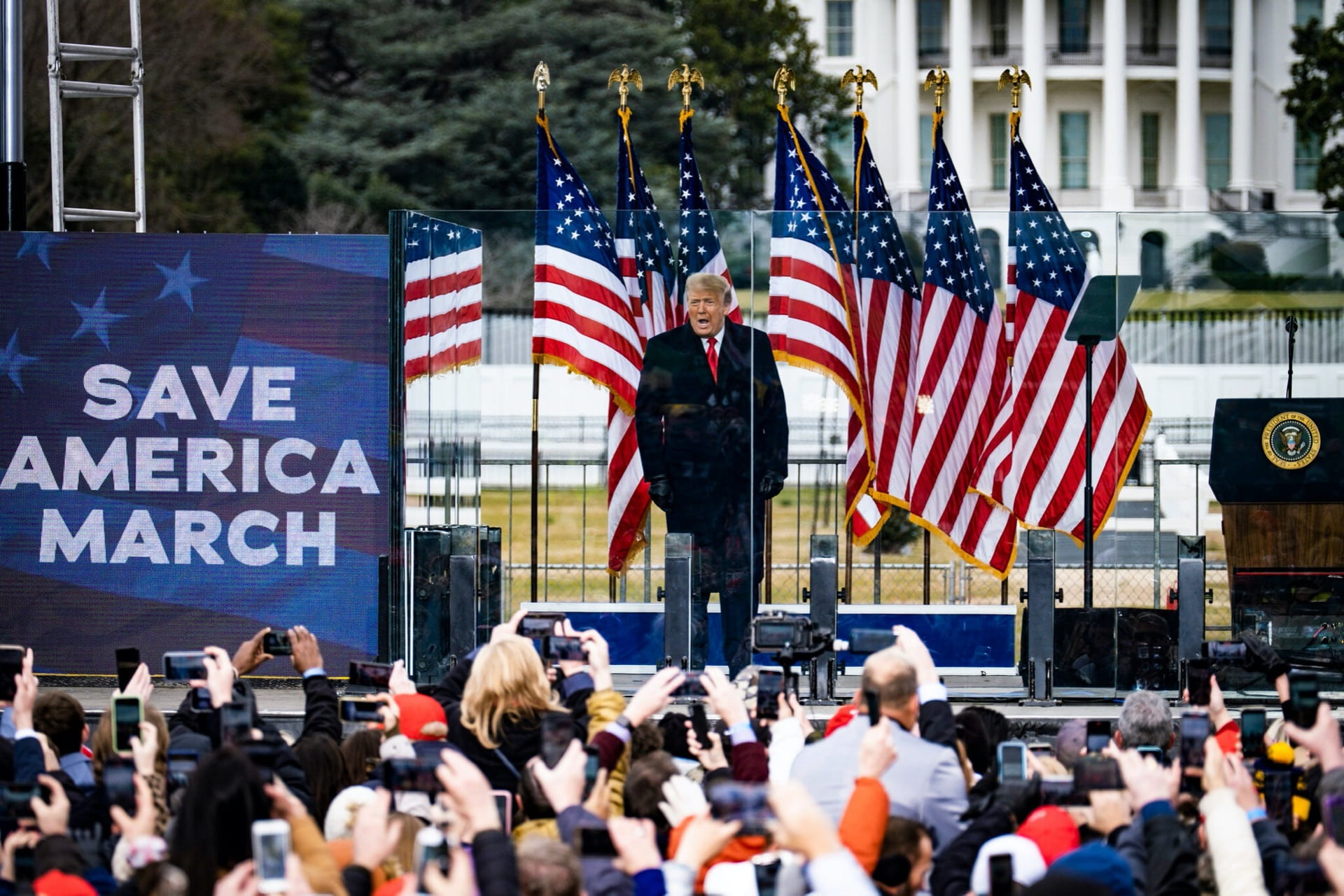 Donald Trump stands on stage next to a sign saying "Save America March" and speaks to a large crowd